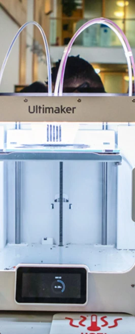 Three Ultimaker 3D printers in a row, with one actively printing an object, in a college workshop setting. Three Ultimaker 3D printers in a row, with one actively printing an object, in a college workshop setting.